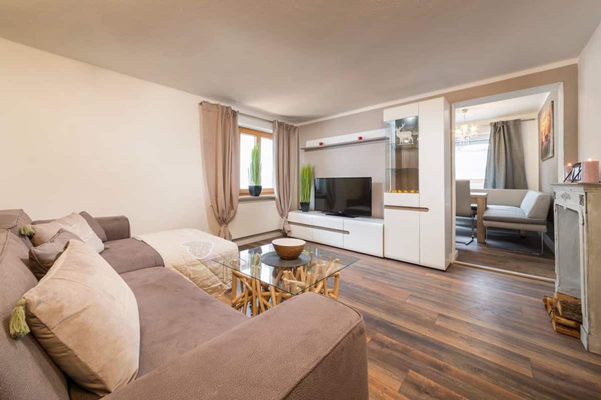 A modern living room with a sofa, a glass coffee table, a flat-screen TV, and a kitchen area with a refrigerator.