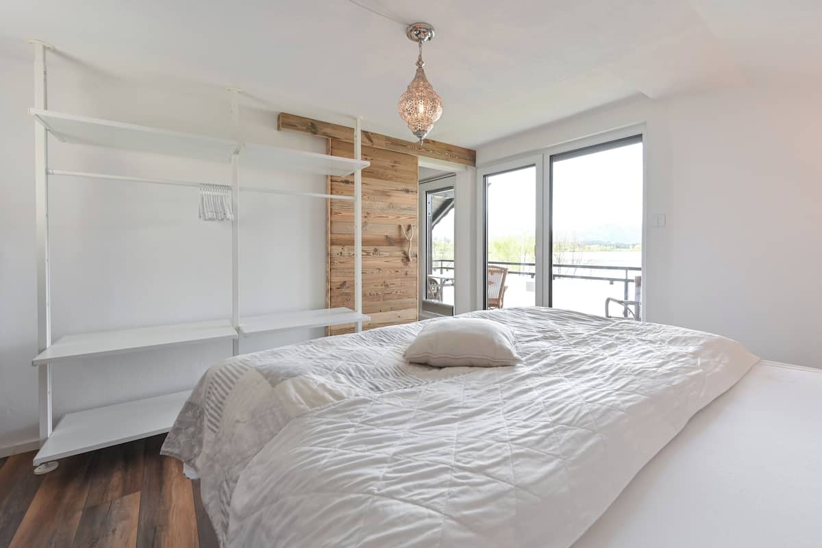 A bedroom with a large bed, built-in wooden shelving, a hanging light fixture, and a view of a balcony.
