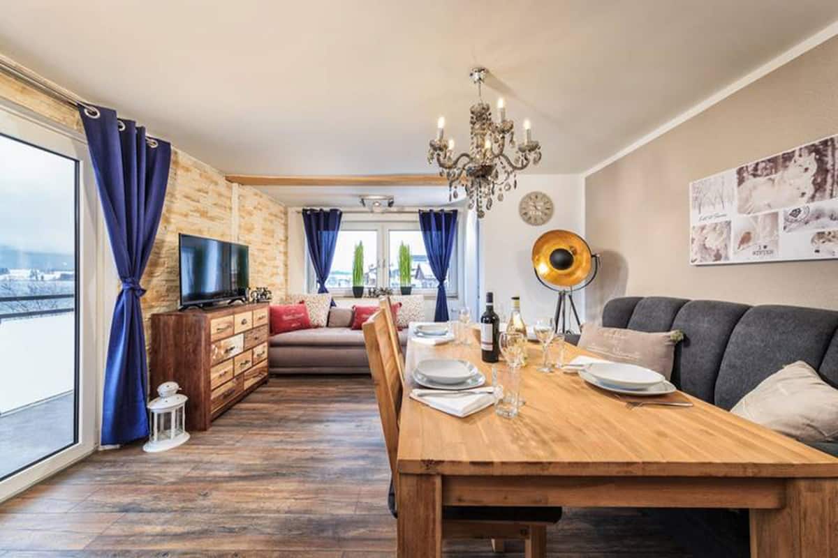 A dining area with a wooden table set for two, a sofa, and a television in the background.