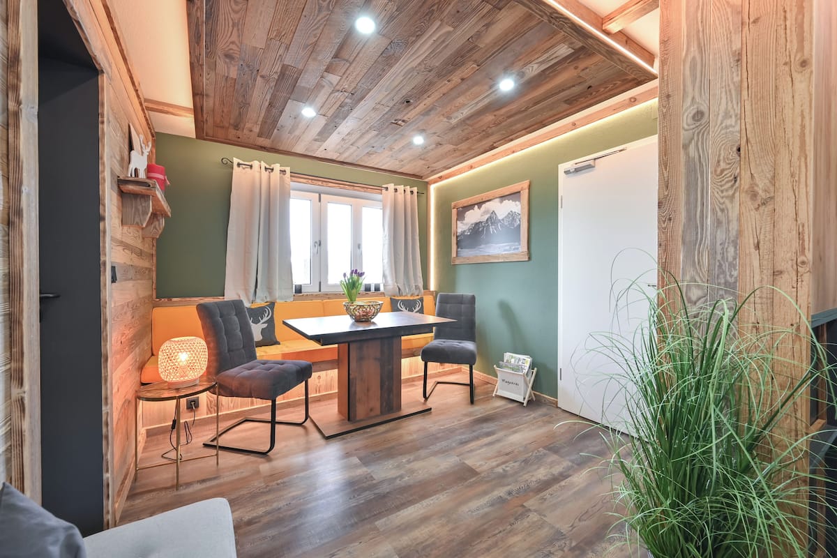 A cozy room with a wooden ceiling, a dining table with chairs, a sofa, a small side table with a lamp, and a large plant.