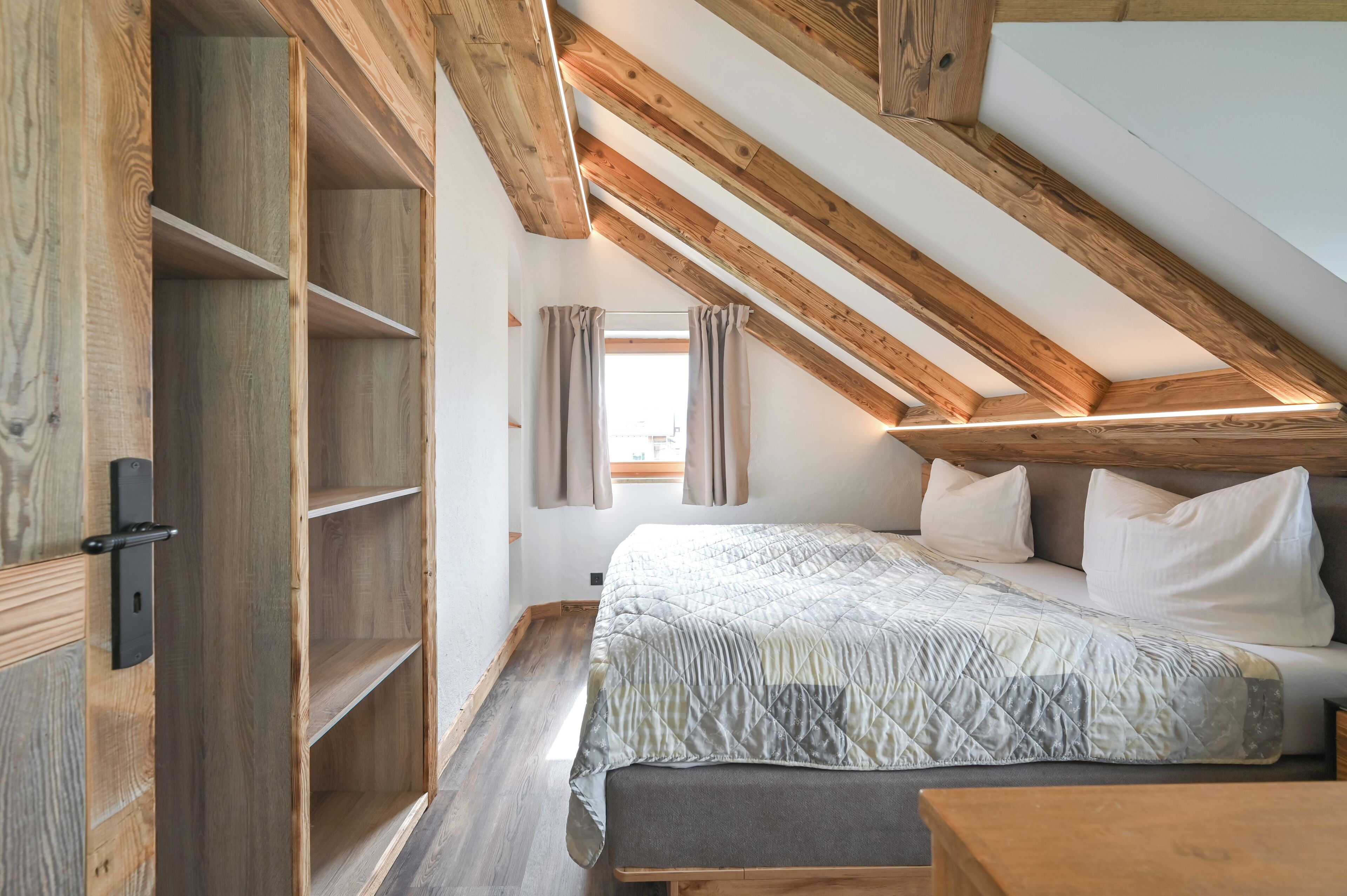 Ein Schlafzimmer mit schrägen Wänden, Holzbalken, einem Bett mit Kissen, einem Nachttisch und einem Fenster mit Vorhängen.