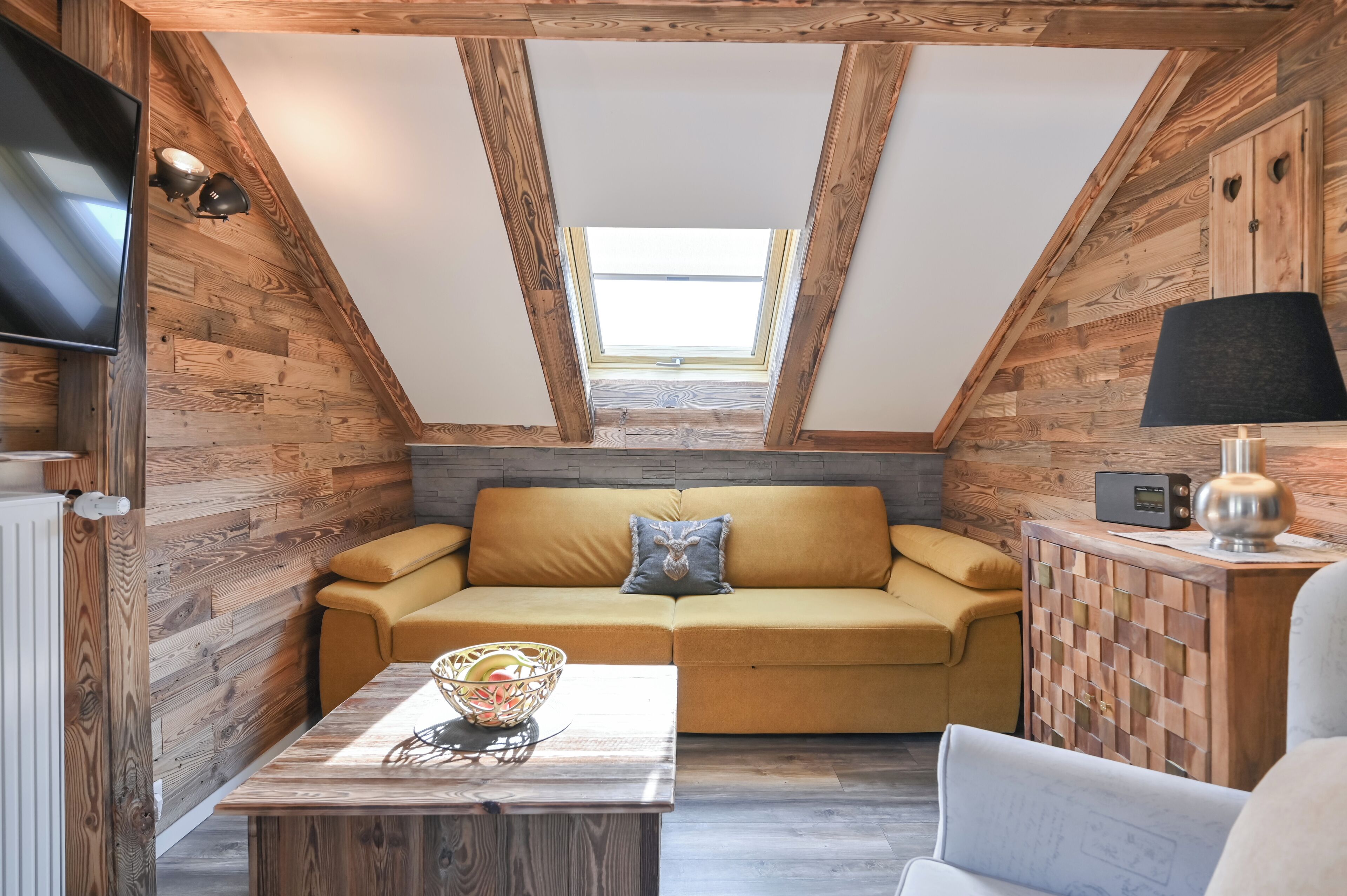 Acogedor dormitorio en el ático con techo de madera, un sofá, una mesa y un televisor.