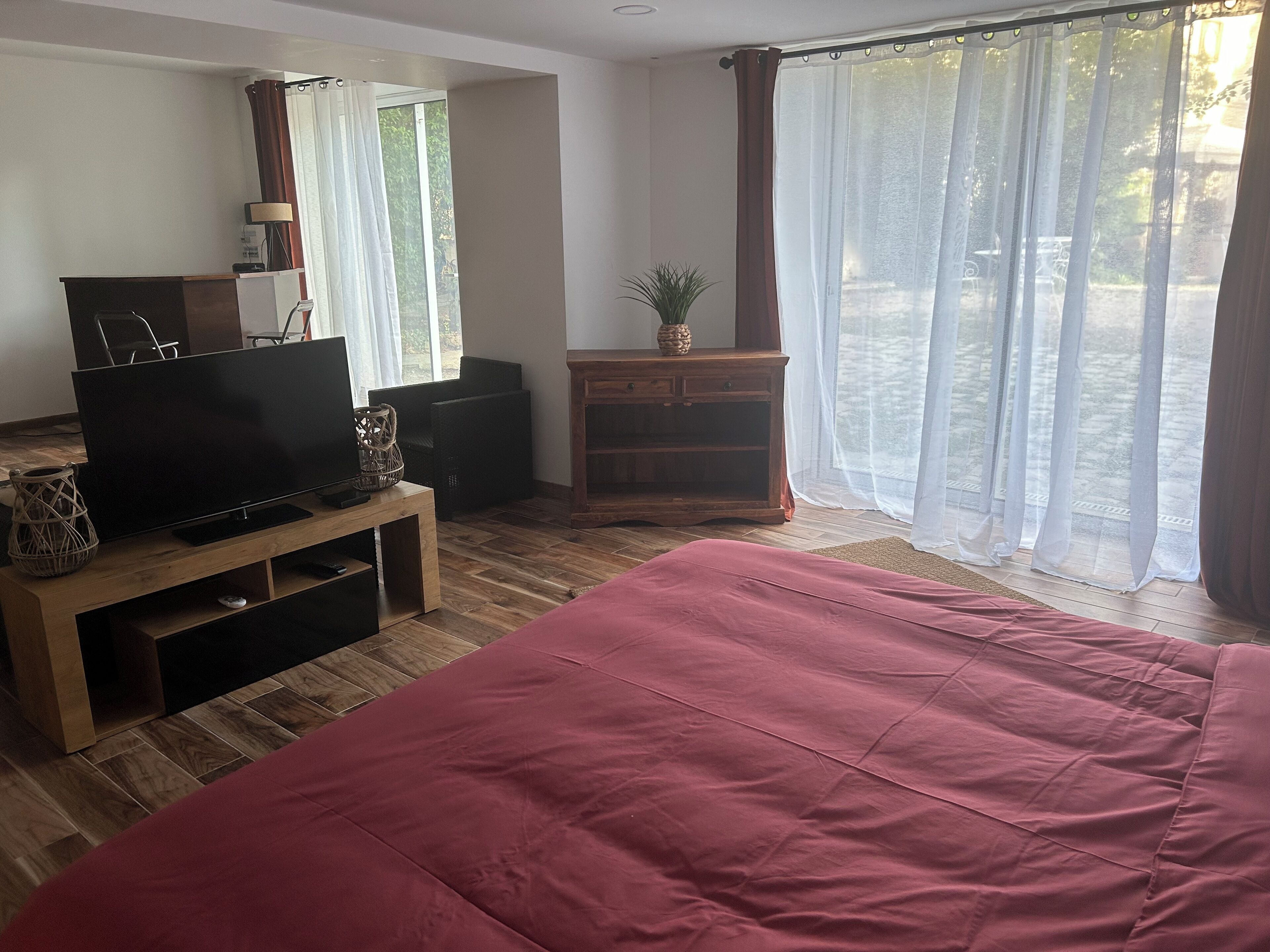 Una habitación con una cama, un televisor sobre un soporte de madera, un pequeño armario de madera y una ventana con cortinas translúcidas.