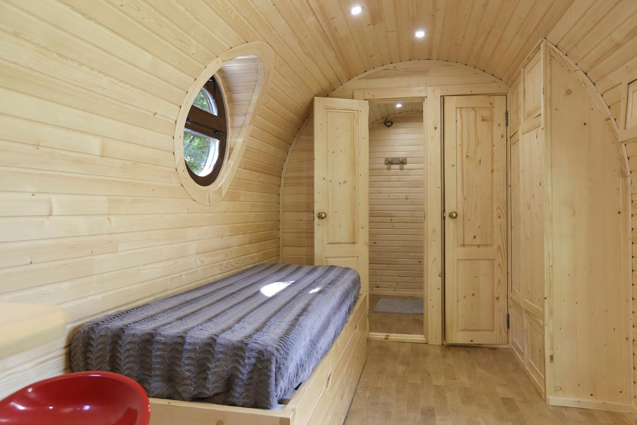 Ein Zimmer in einer Holzhütte mit einem Bett, einem kleinen Fenster und einer Tür zu einem weiteren Raum.