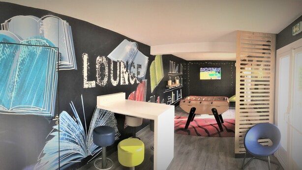 A modern room with a black wall featuring large book illustrations and the word "LOUNCE". There is a white desk, a blue chair, and a yellow stool. A television is mounted on the wall, and a wooden slat partition is visible. - Hoteleco Sud (Le Passage)
