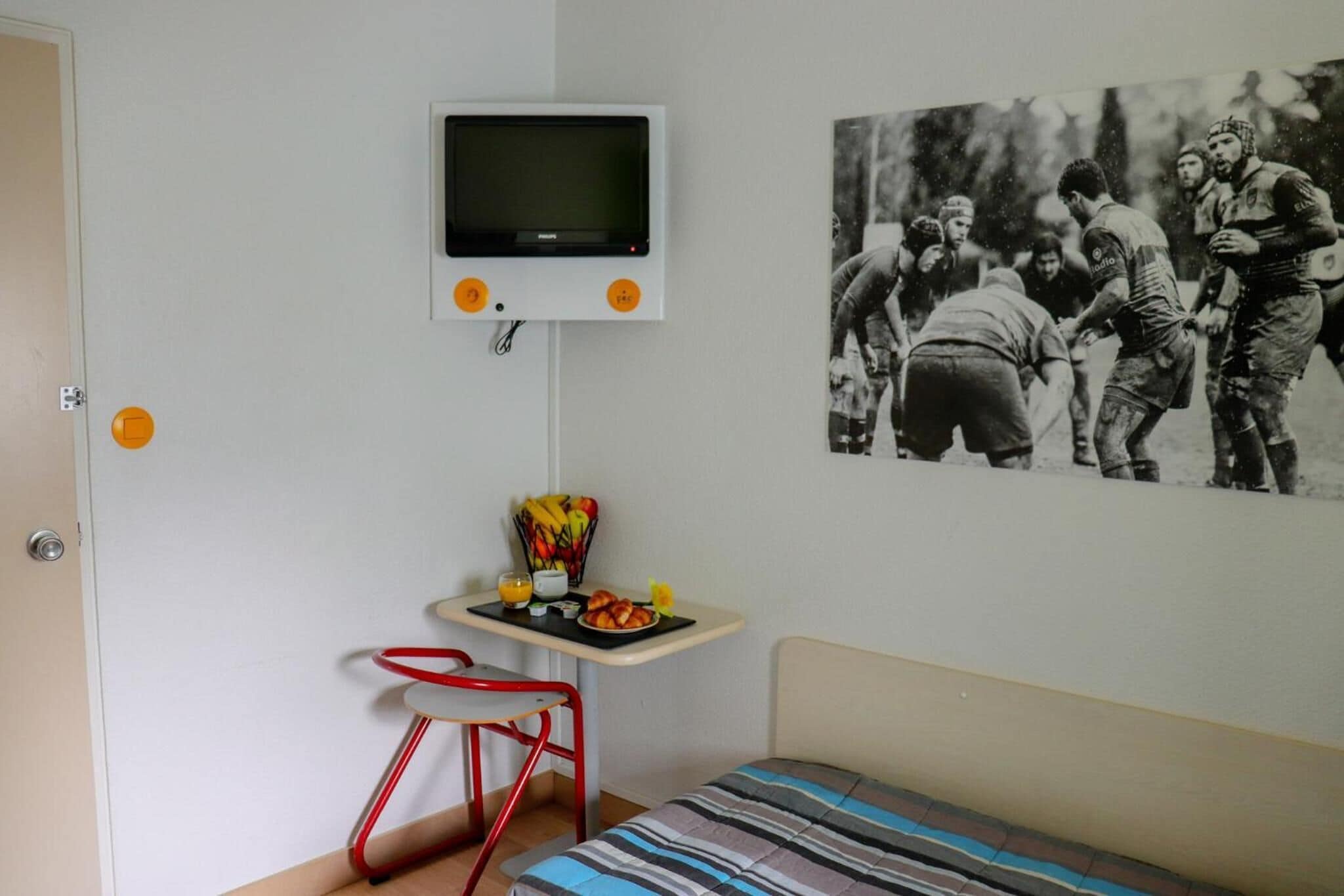 Una habitación con una cama, una mesita con una cesta de frutas, un televisor montado en la pared y una foto enmarcada en la pared.