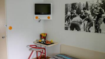 Una habitación con una cama, una mesita con una cesta de frutas, un televisor montado en la pared y una foto enmarcada en la pared.