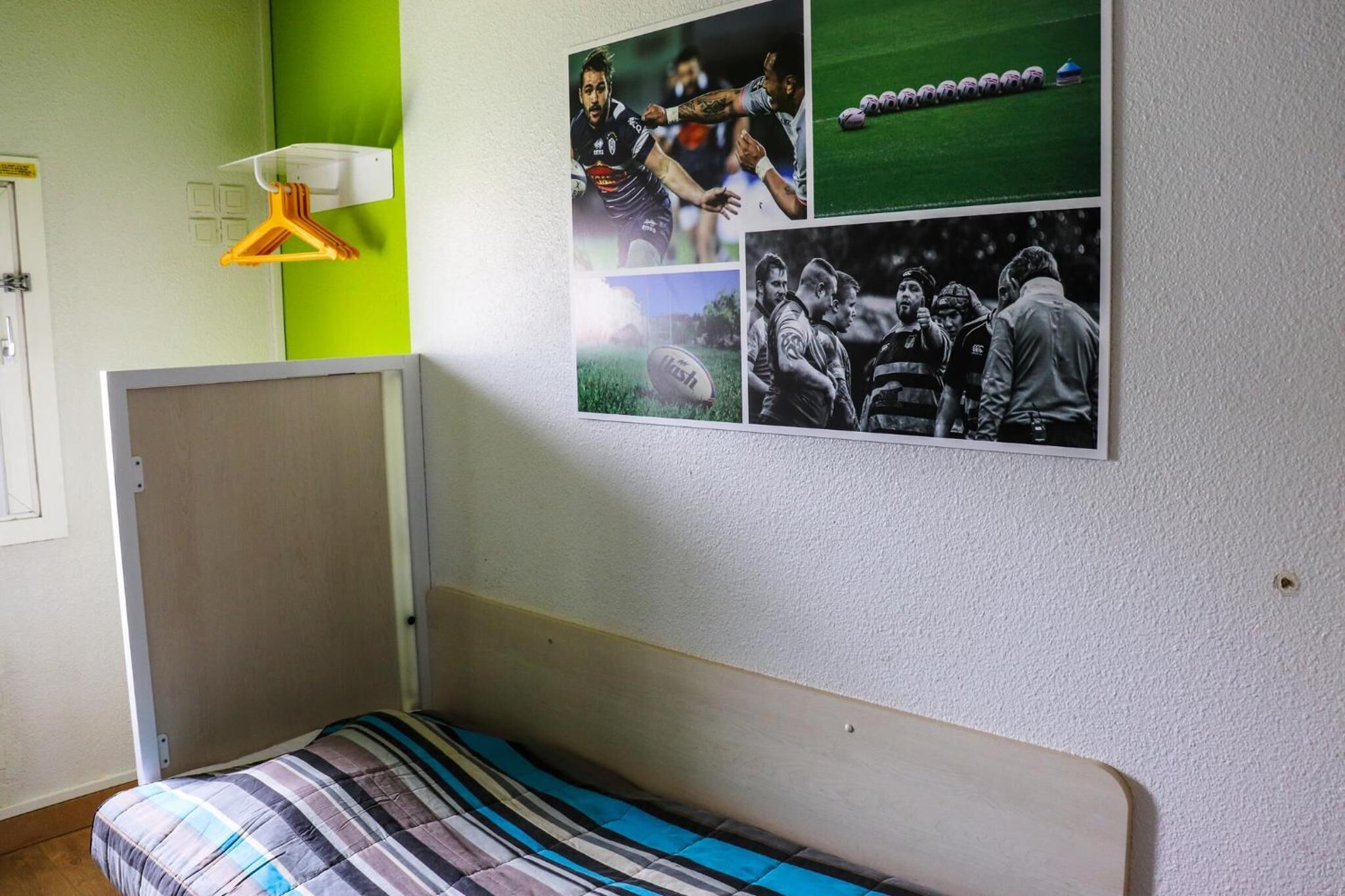 Una habitación con una cama, un estante en la pared con una lámpara y un tablón de anuncios con fotos deportivas.