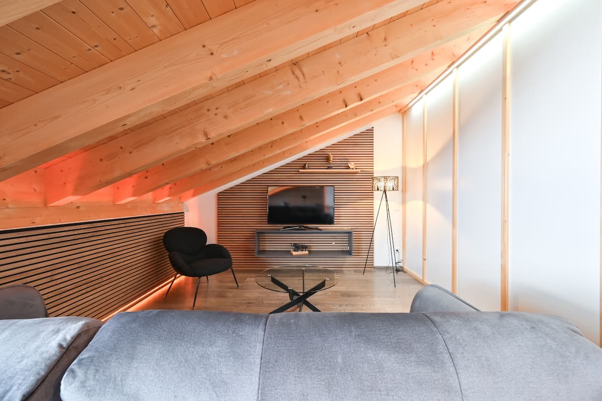 A modern loft bedroom with a sloped ceiling, a large gray sofa, a black chair, a glass coffee table, and a TV mounted on a wooden wall.