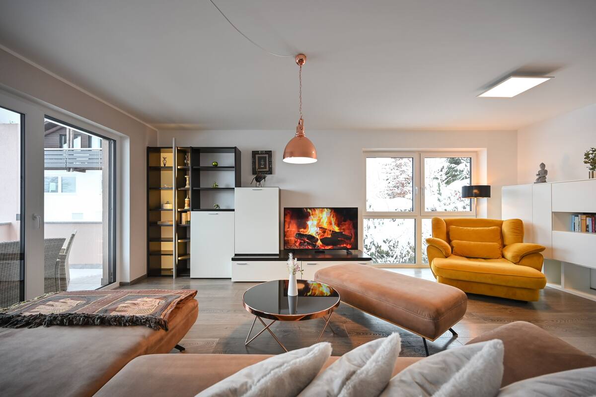 A modern living room with a sofa, ottoman, and a round coffee table. There is a television and a fireplace in the background.