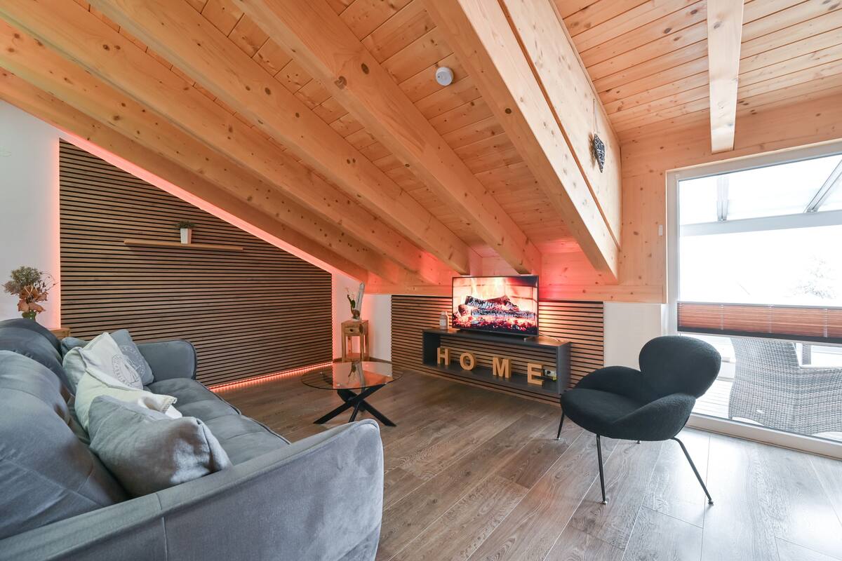 A modern living room with a wooden ceiling, a grey sofa, a black armchair, a glass coffee table, and a TV on a wooden stand.