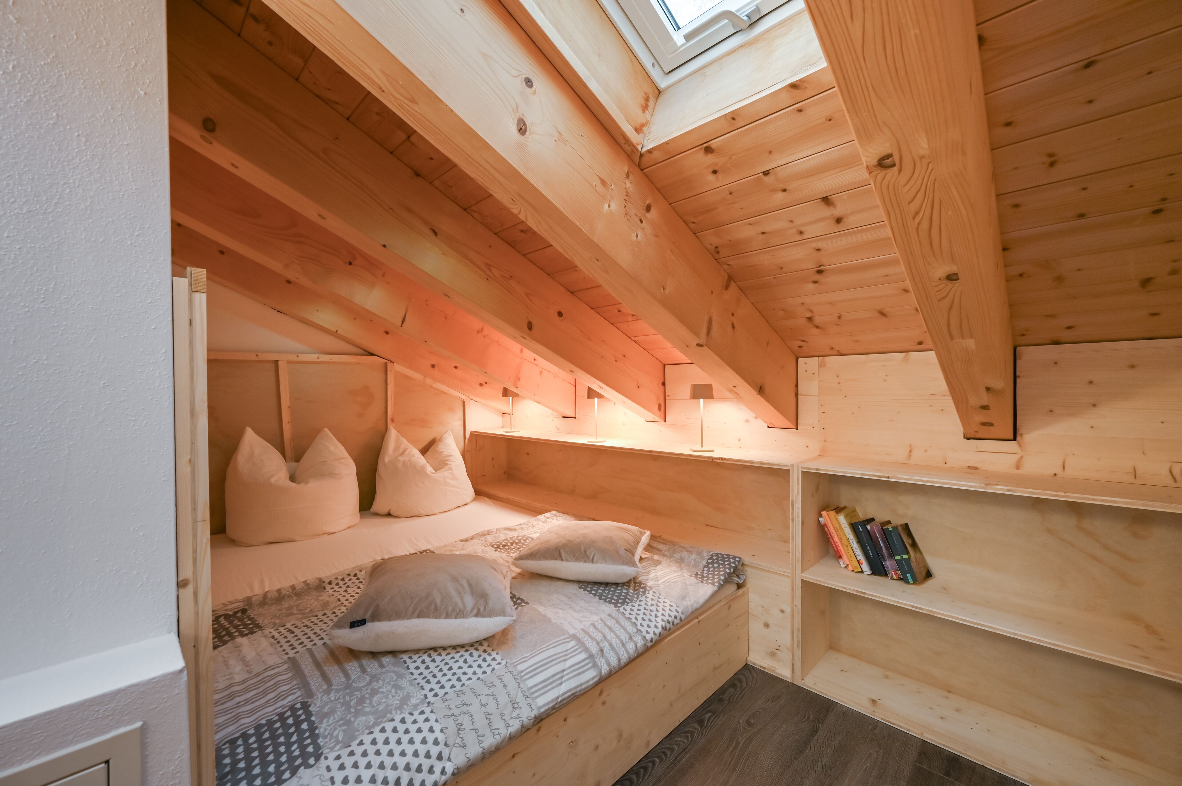 Una camera da letto mansardata accogliente, con soffitto spiovente, letto a scomparsa e una mensola piena di libri.