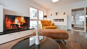 Una sala de estar moderna con un televisor de pantalla plana, un sillón amarillo, un aparador blanco con libros y un jarrón con flores sobre una mesa de centro.