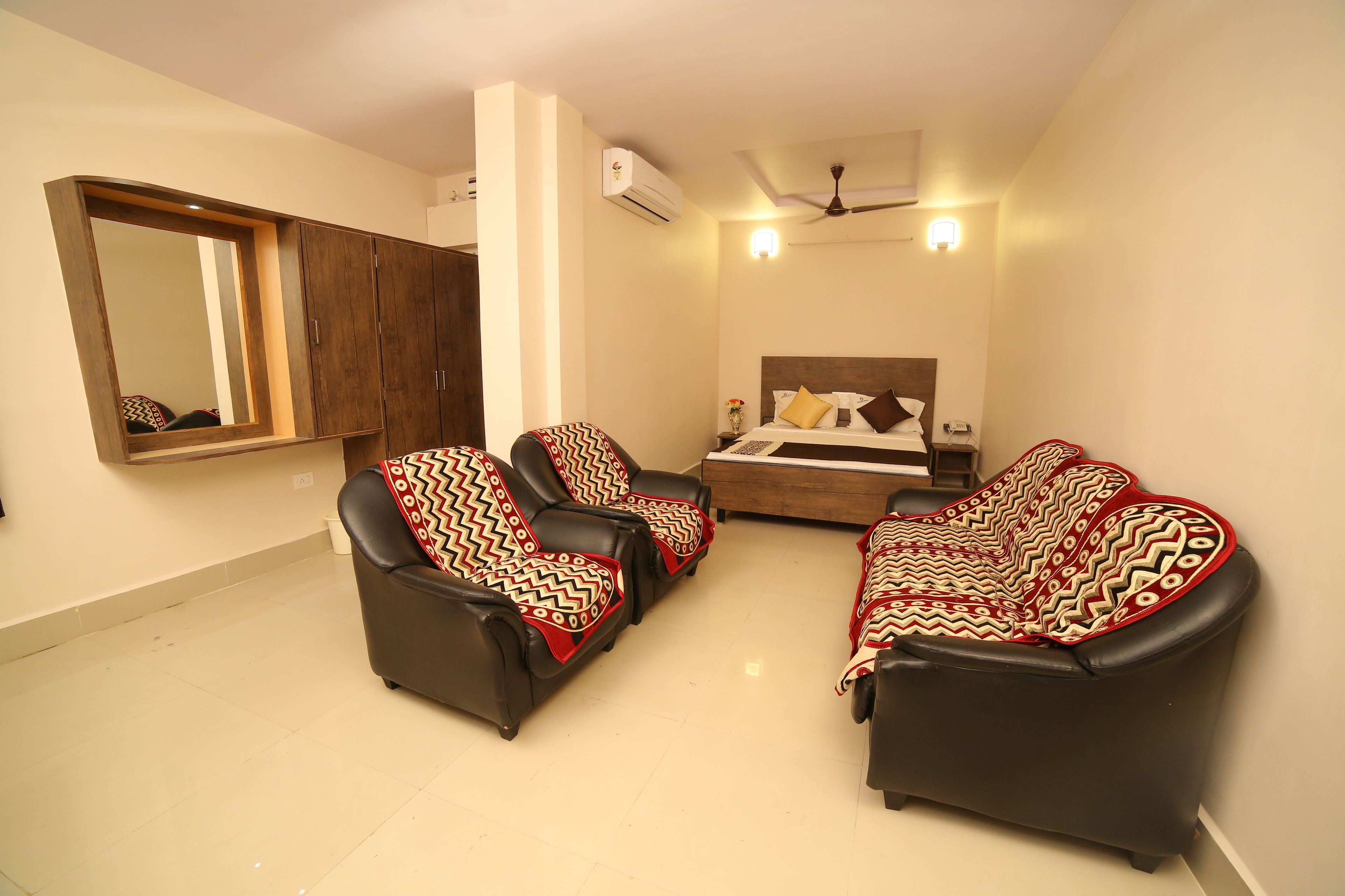 A compact hotel room with a bed, a sofa, a chair, a large mirror, and a ceiling fan.