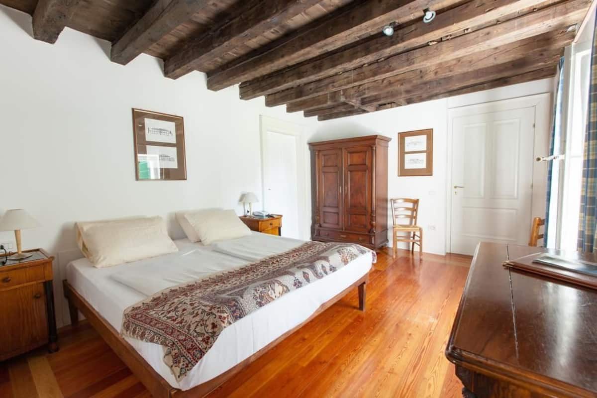 A bedroom with a large bed, wooden floor, and a ceiling with exposed beams. There is a wardrobe, a chair, and a desk with a lamp.