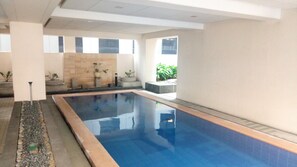 Uma piscina interior com água cristalina, rodeada por piso e paredes azulejadas, com plantas em vasos e uma parede de pedra ao fundo.