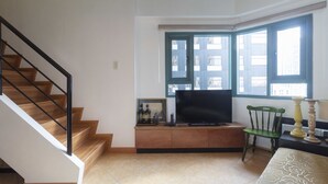 Un soggiorno con una scala, una televisione su un mobile in legno, una poltrona verde e una finestra con persiane.