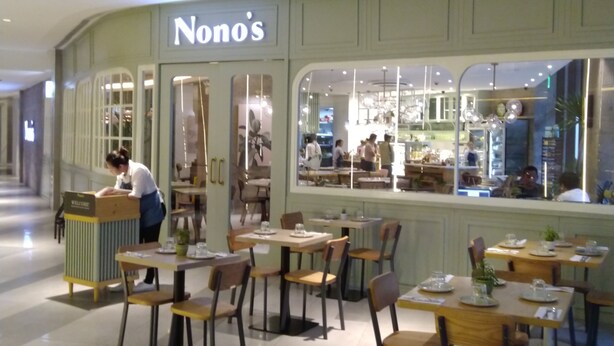 A restaurant named 'Nono's' with a modern interior, including wooden tables and chairs, and a welcoming staff member at the entrance.
