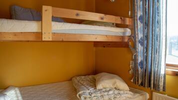 Una litera con una cama inferior y una cama individual debajo. Hay una ventana con cortinas, un radiador y una lámpara fijada a la pared.