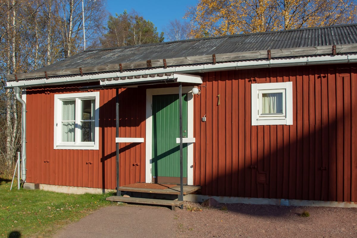 A red wooden building with a green door, a small window, and a metal roof.