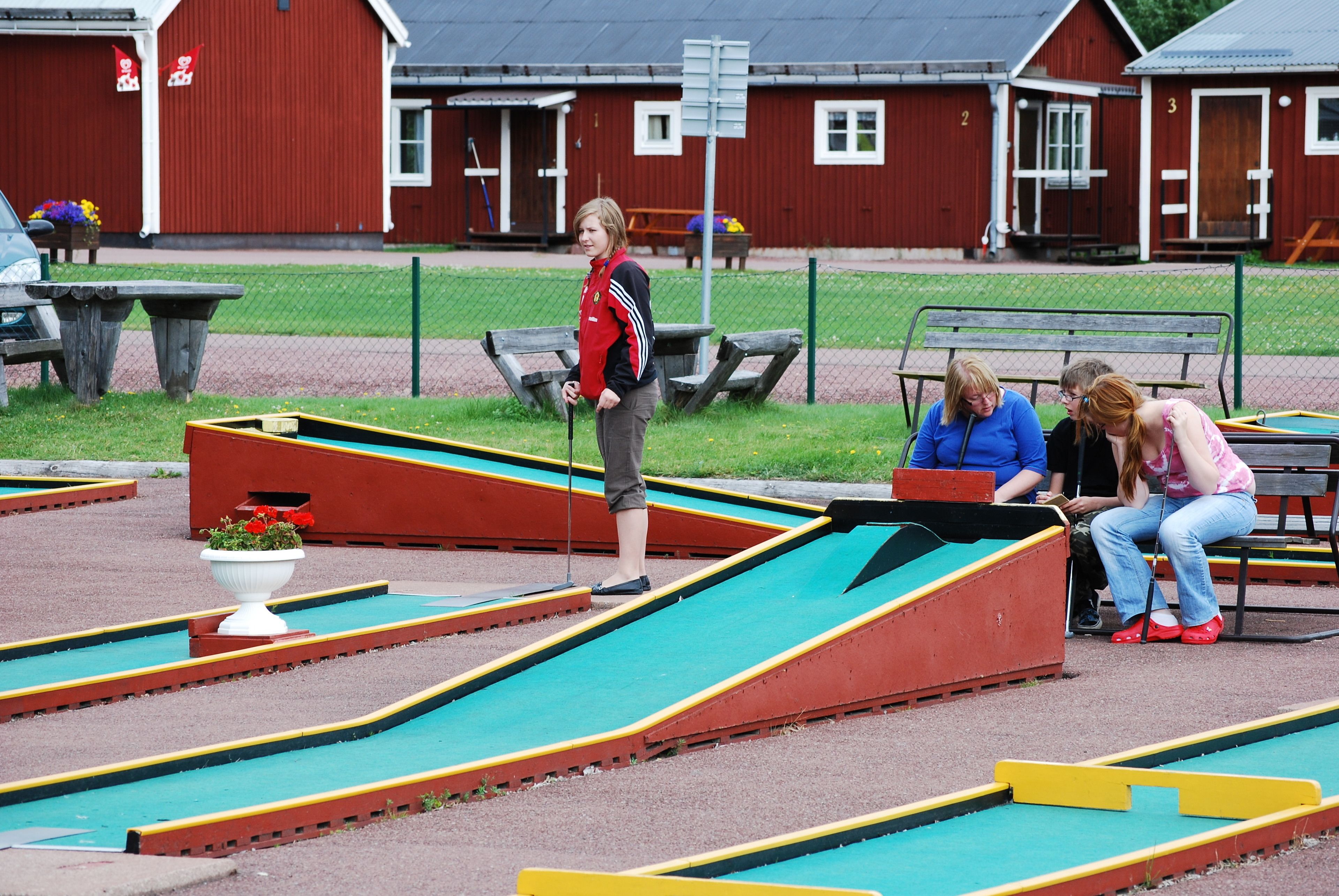 En miniatyrgolfbana med gröna och röda ramper, en vit blomkruka och en röd byggnad i bakgrunden.