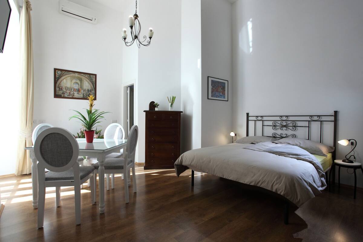 A bedroom with a bed, bedside table, lamp, and a dining area with chairs and a table.