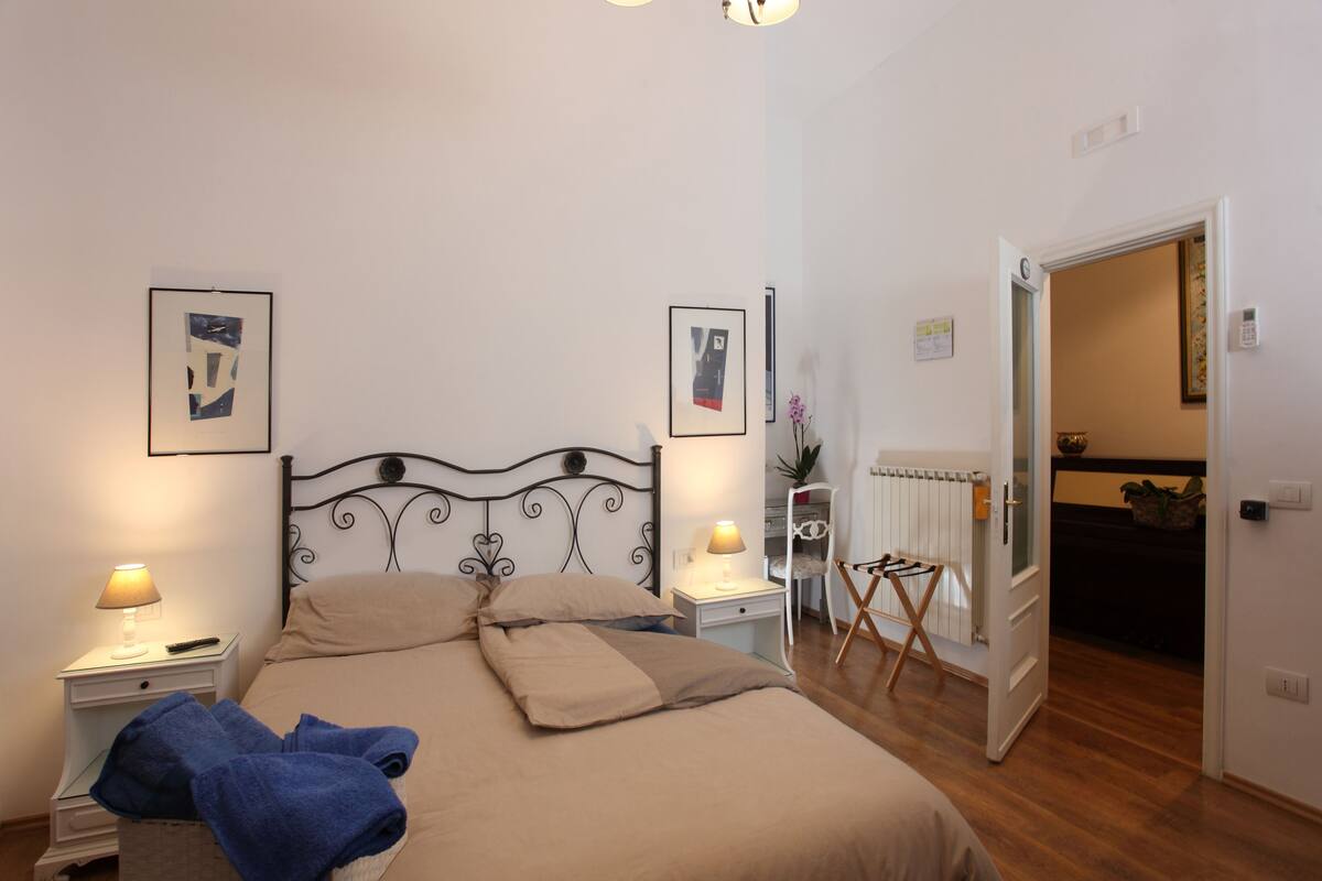 A bedroom with a bed, two bedside tables with lamps, a radiator, and a door leading to another room.
