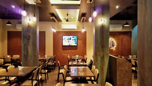 Um restaurante com várias mesas e cadeiras, uma televisão presa à parede e ventiladores de teto.