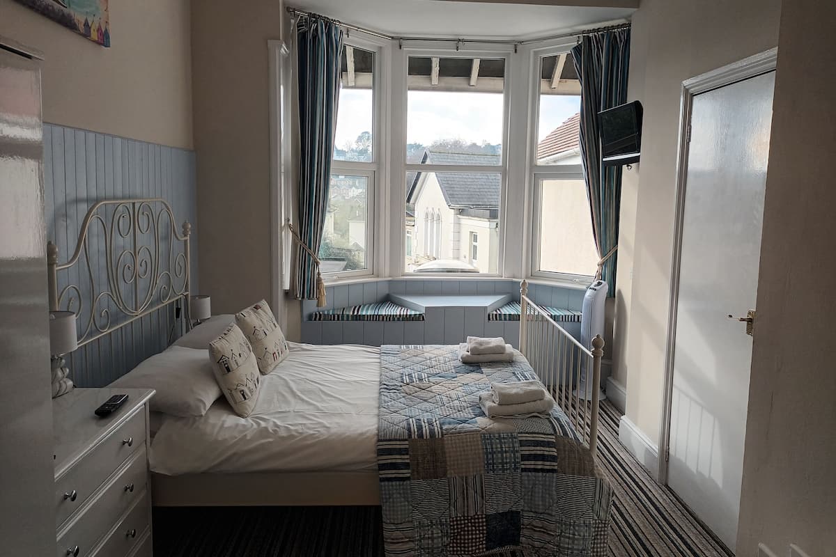 A bedroom with a bed, a dresser, a window seat, and a view of buildings outside.