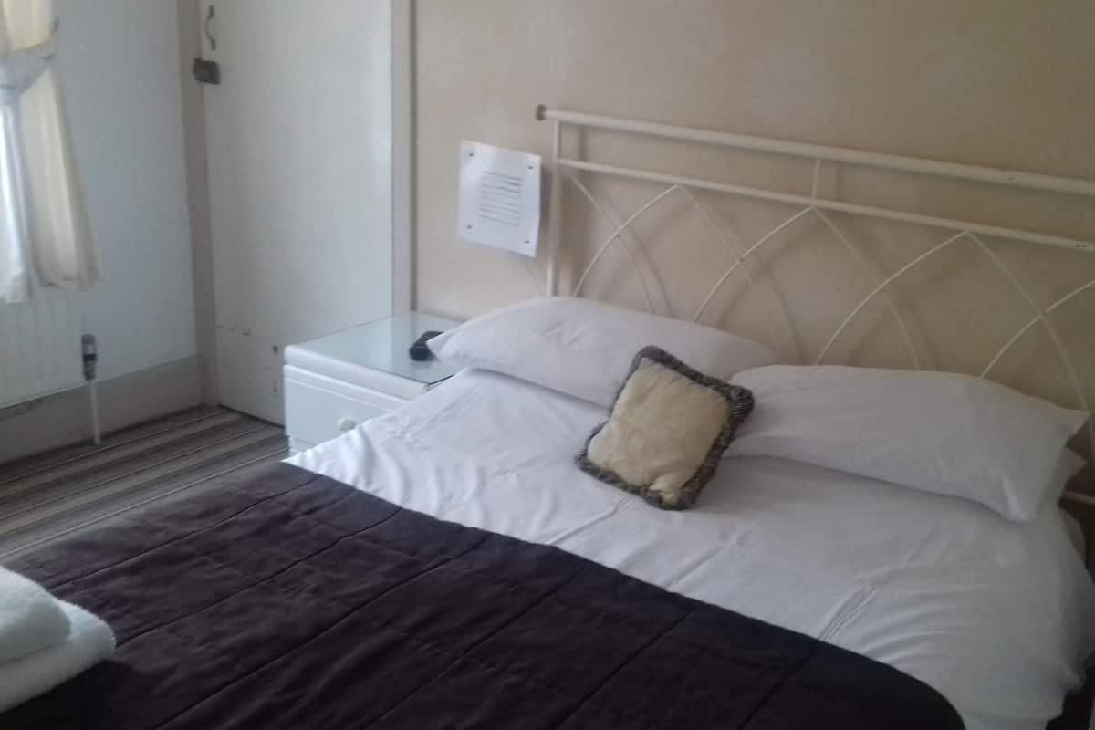 A neatly made bed with a dark-colored blanket, a white pillow, and a headboard with a paper attached. There is a small bedside table with a lamp and a power outlet.