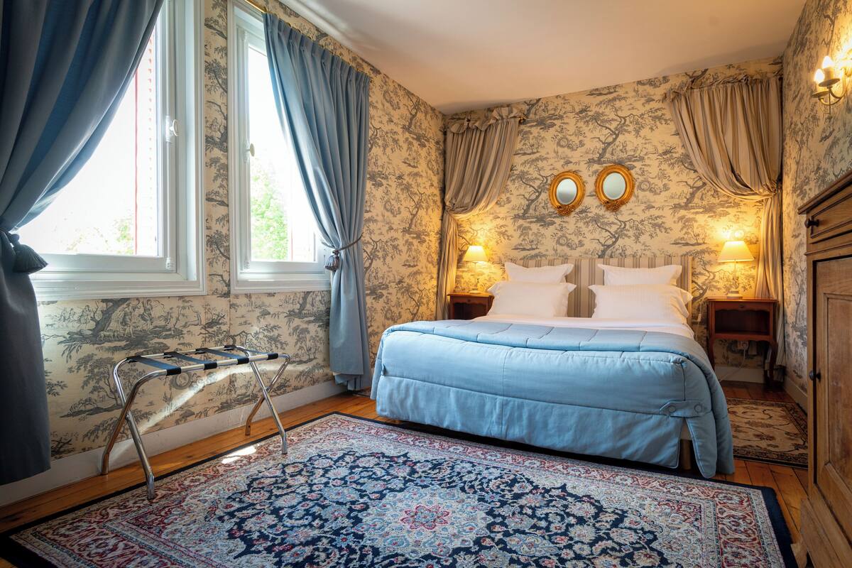 Een slaapkamer met een bed, twee bedlampjes, een nachtkastje, een raam met gordijnen, een houten vloer en een kamerkleed met een patroon.
