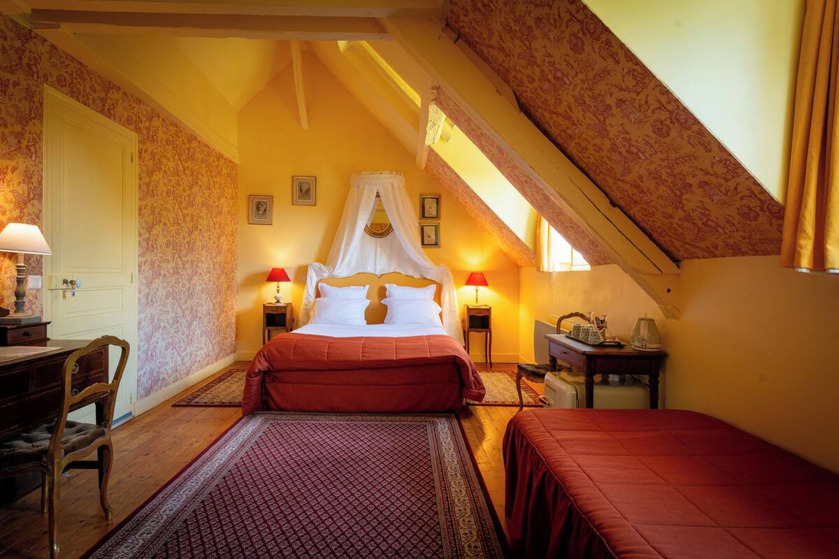 Een slaapkamer met een groot bed, een bureau en een stoel. Er is een raam met gordijnen en een lamp op het bureau.
