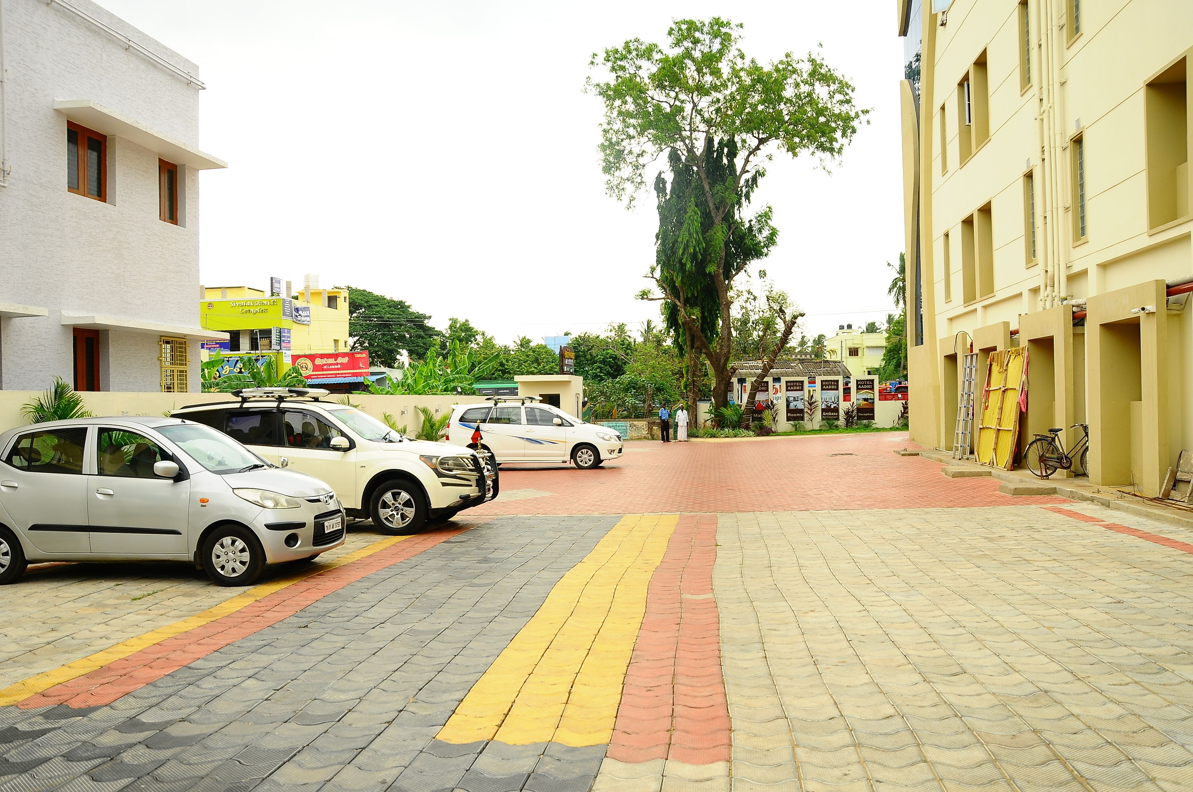 Un área de estacionamiento con varios coches, un árbol y edificios al fondo.