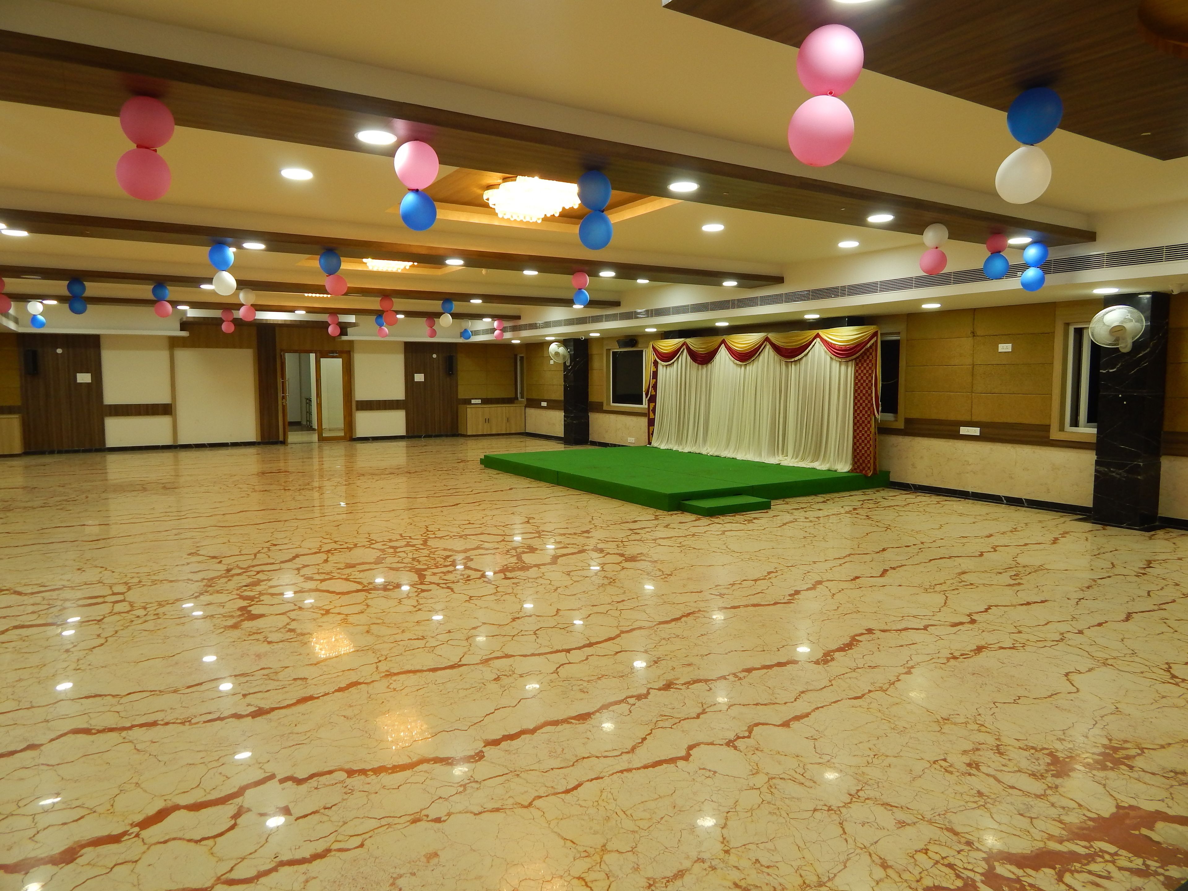 Ein geräumiger Saal mit poliertem Marmorboden, einer Bühne mit grünem Teppich und einer Decke, die mit bunten Ballons verziert ist.