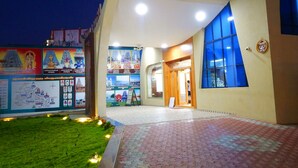 A building with a tiled entrance, a grassy area, and a poster with images and text. - Hotel Aadhi (Mayiladuthurai)