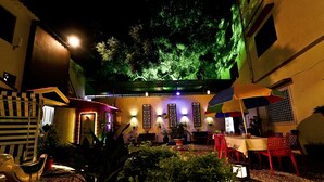 An outdoor dining area with tables and chairs, potted plants, and a pergola with string lights. - Hotel Royal (Diu)