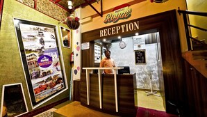 A reception area with a sign reading "Hotel Royal Reception," a man standing behind the counter, and a large framed poster on the wall. - Hotel Royal (Diu)