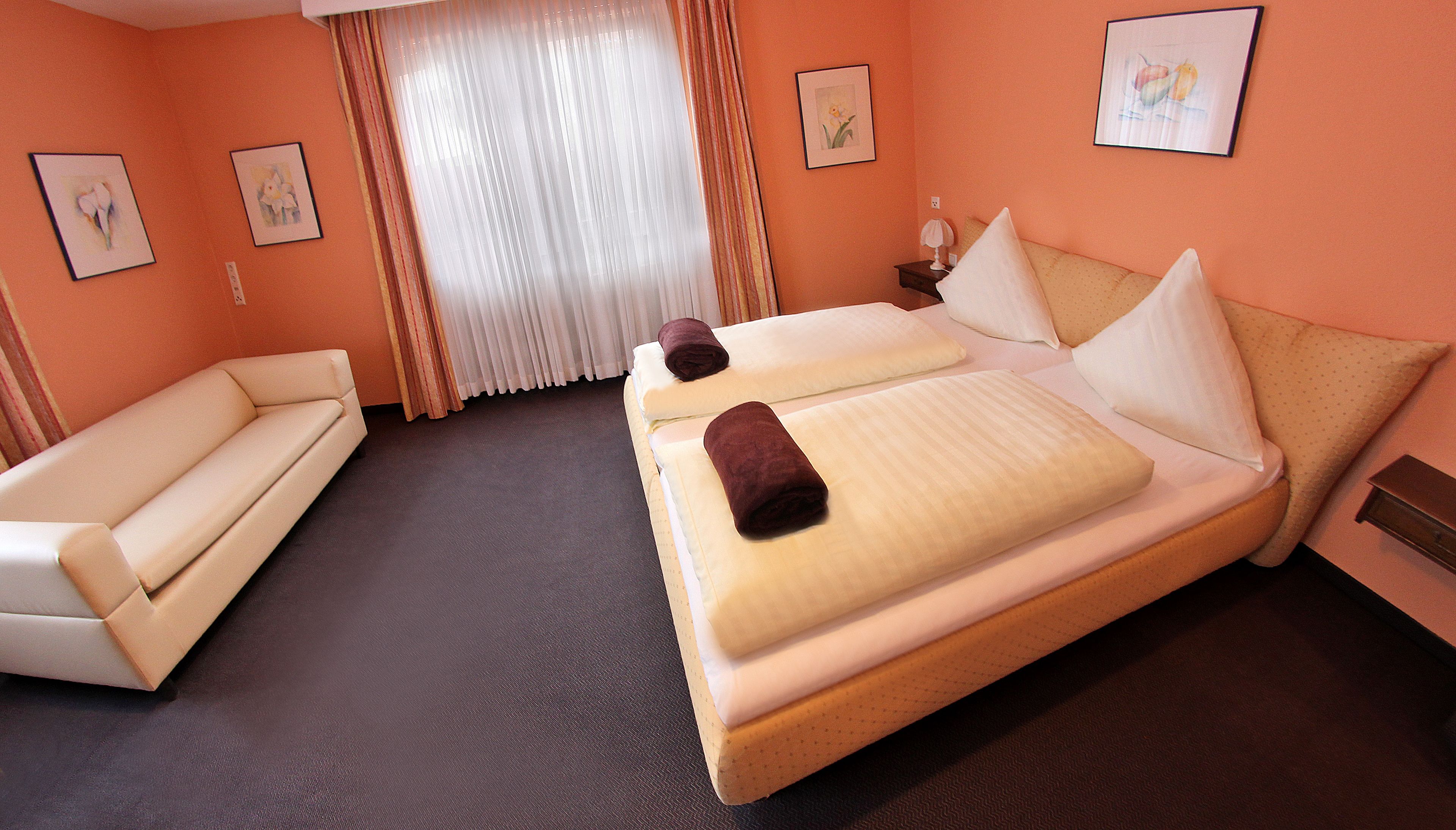 A hotel room with a double bed, a sofa, a nightstand with a lamp, and two framed pictures on the wall.