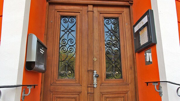 Uma porta dupla de madeira com painéis de vidro e puxador de metal, embutida num edifício com paredes laranja e branco.