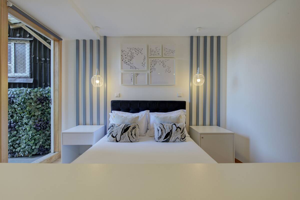 A modern bedroom with a large bed, striped wall panels, and a view of greenery outside.