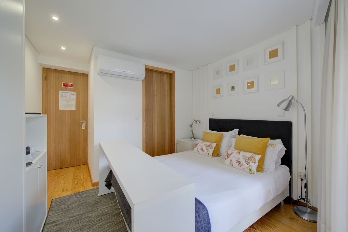A modern hotel room with a bed, bedside table, lamp, and a small desk with a chair.