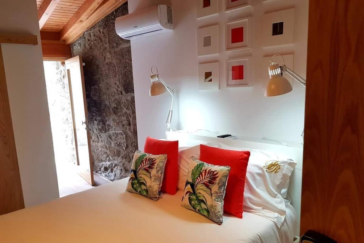 A bedroom with a bed, two red pillows, a white bedspread, a wall-mounted air conditioner, and a bedside lamp.