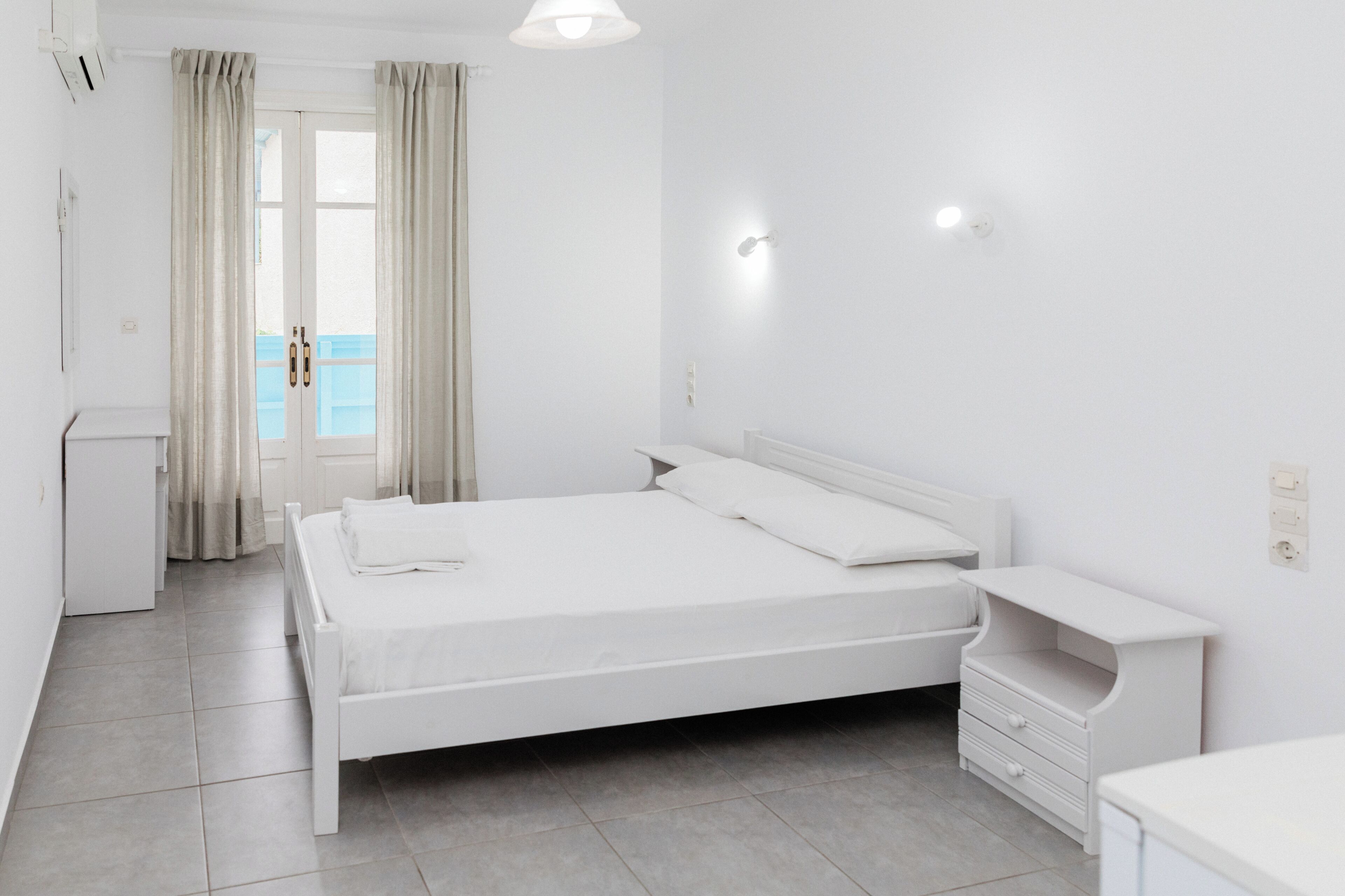 A white room with a bed, a small desk, and a window with curtains.
