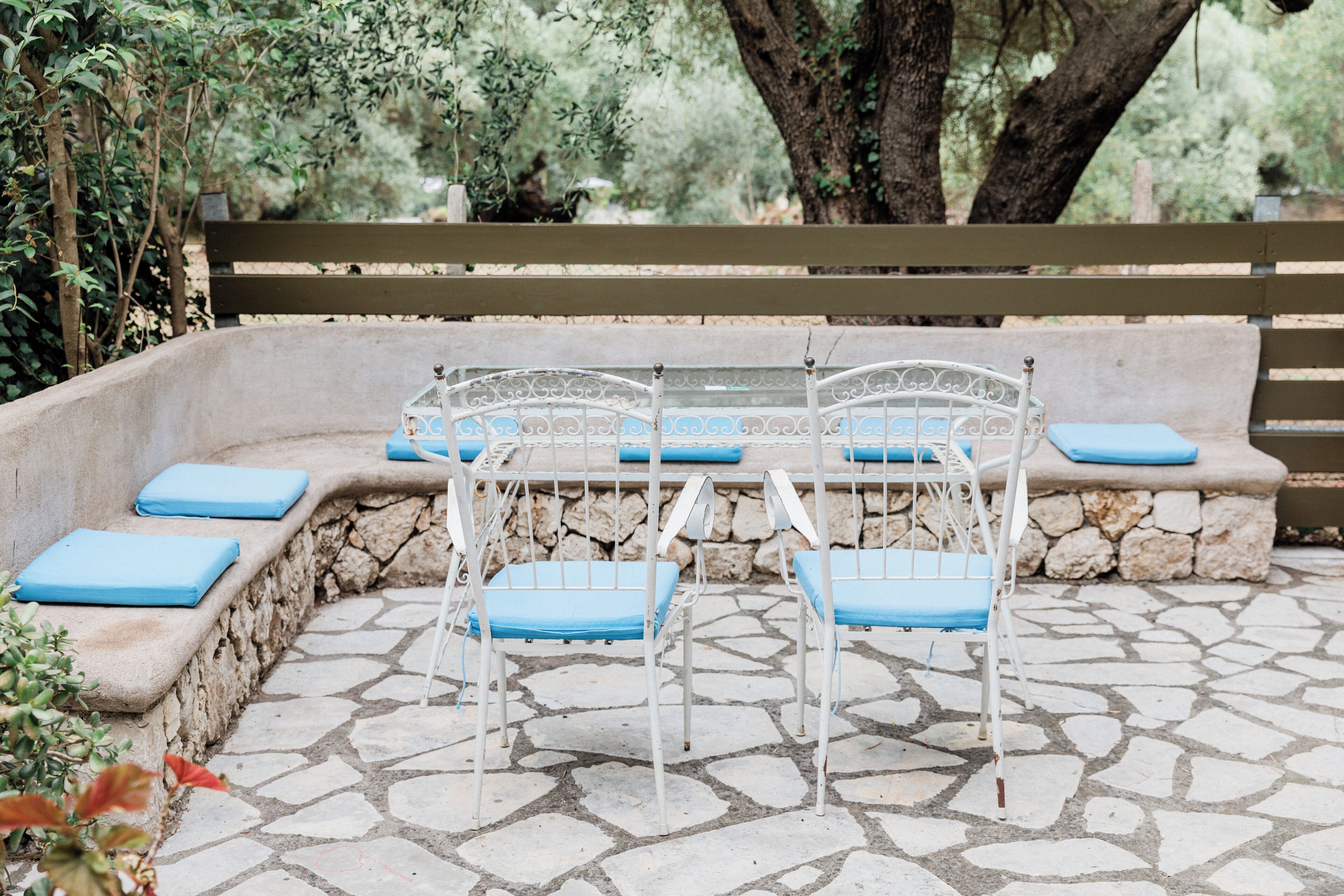 A stone bench with blue cushions, a white metal table, and a wooden bench in a garden setting.