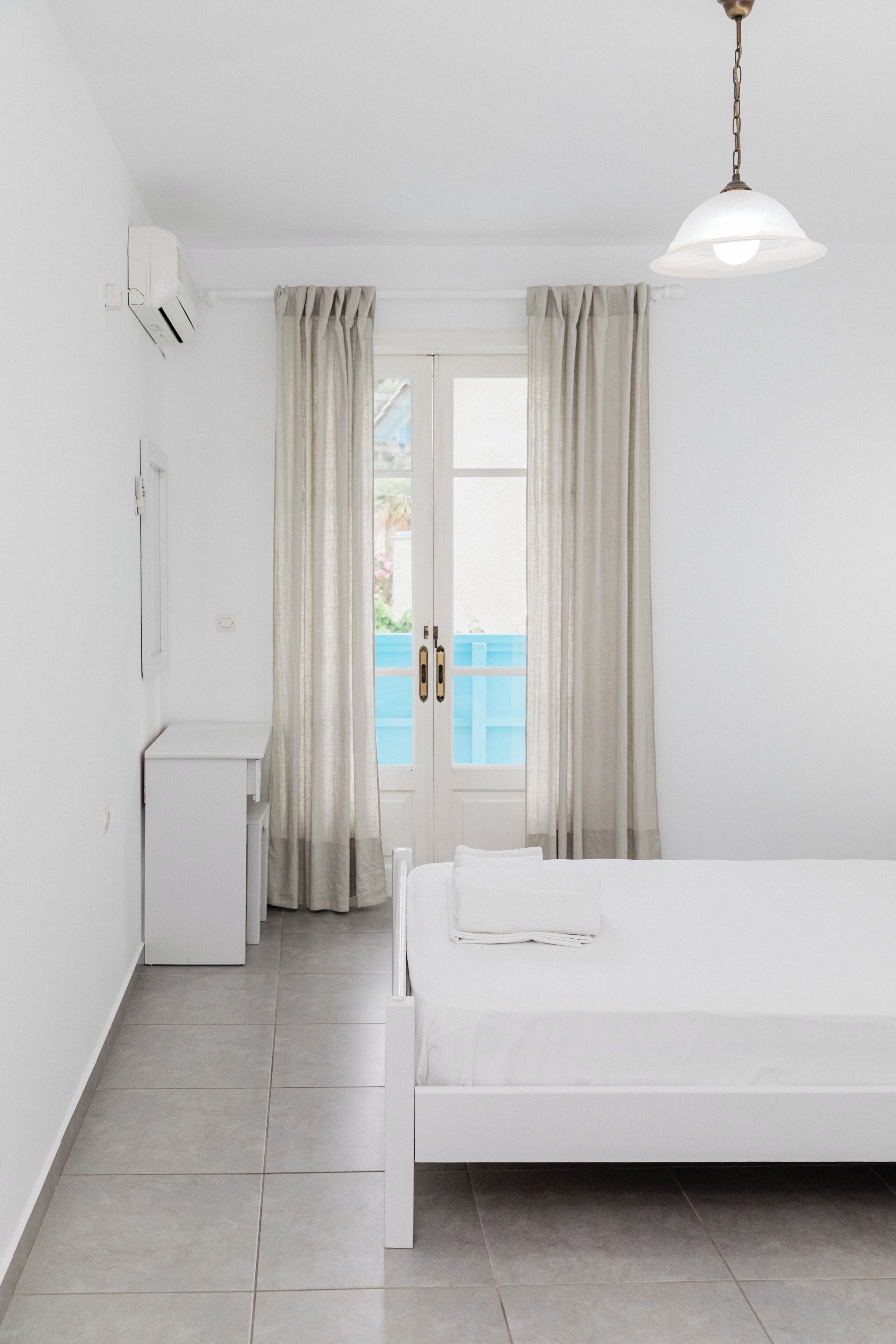 Habitación de hotel minimalista con cama, escritorio pequeño y ventana con cortinas.
