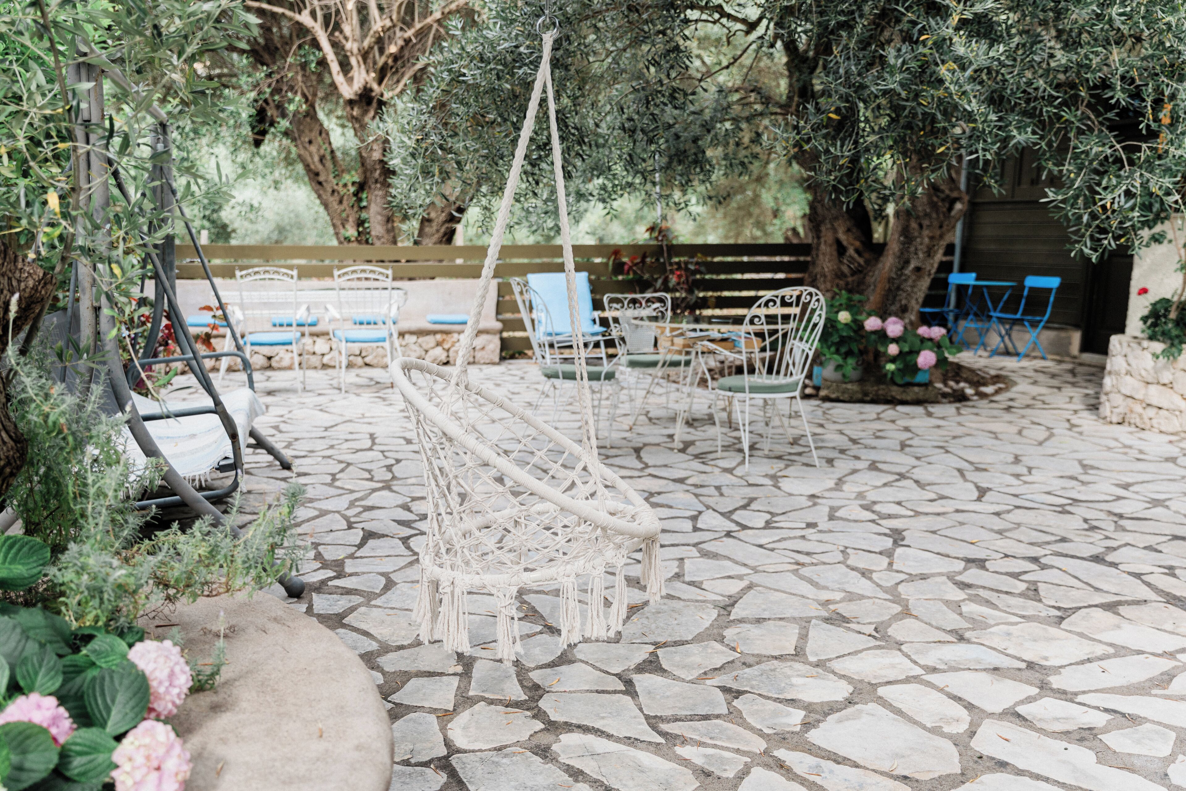 A stone patio with outdoor seating, including white chairs and a hanging chair, surrounded by greenery and trees.