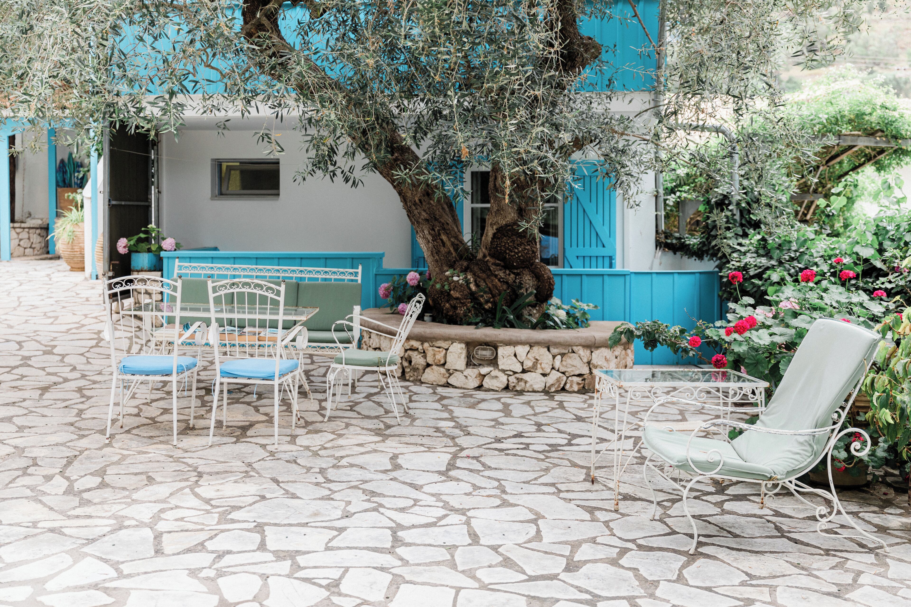 An outdoor seating area with stone flooring, white metal chairs, and a large tree.
