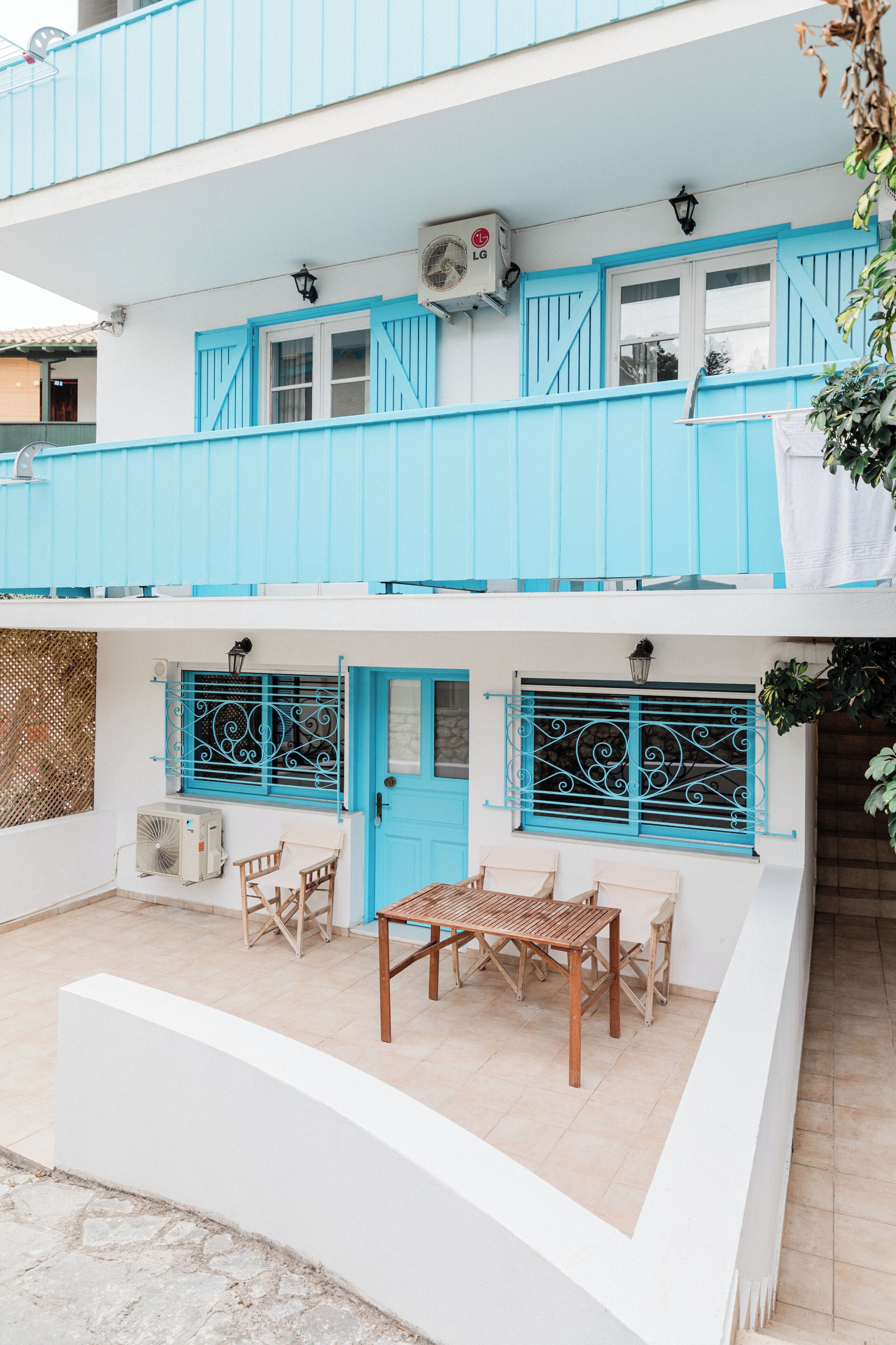 A two-story building with a blue exterior, a balcony with a table and chairs, and an air conditioning unit.