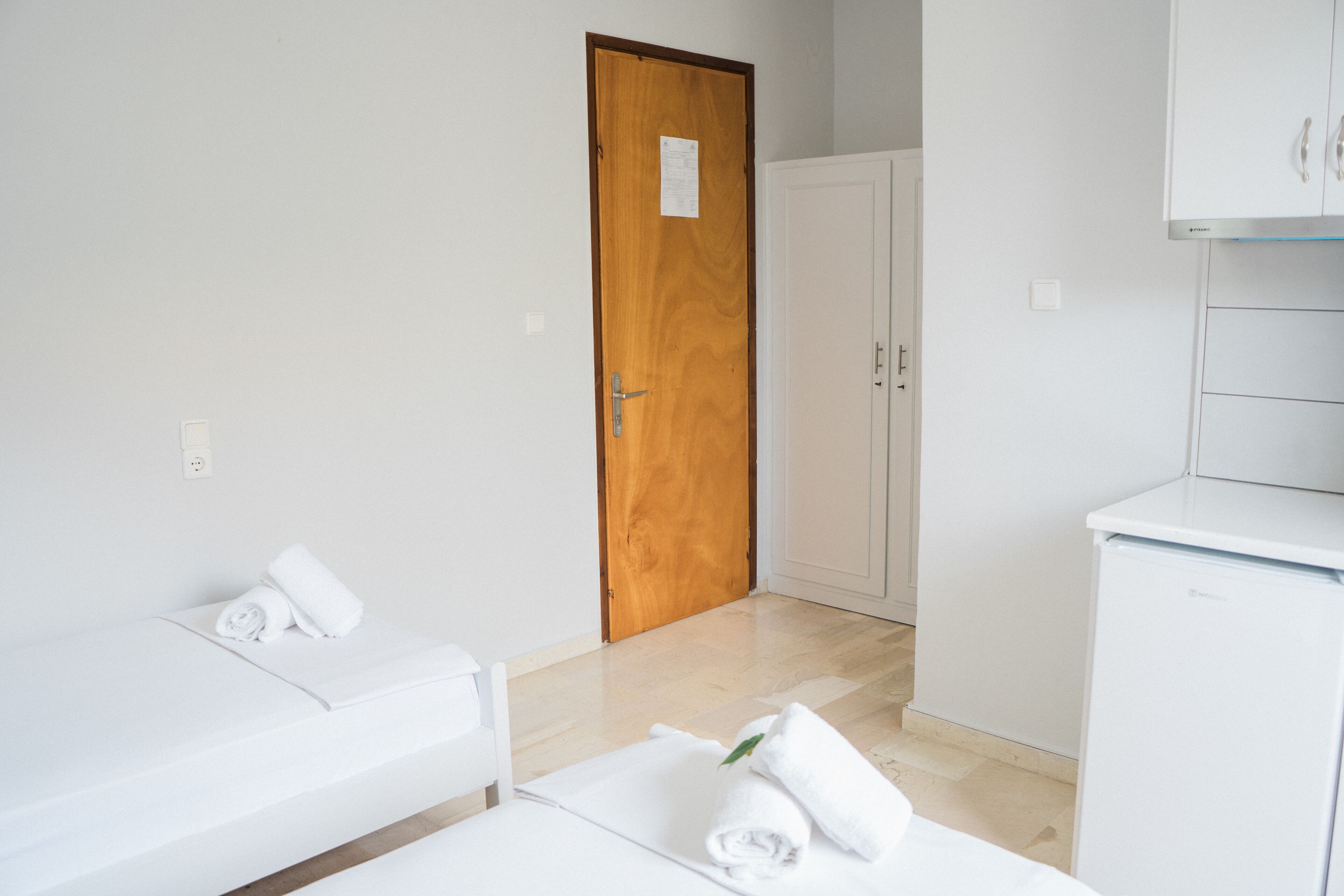A hotel room with a wooden door, a white wardrobe, a white bed with a rolled white towel, and a white bedside table.
