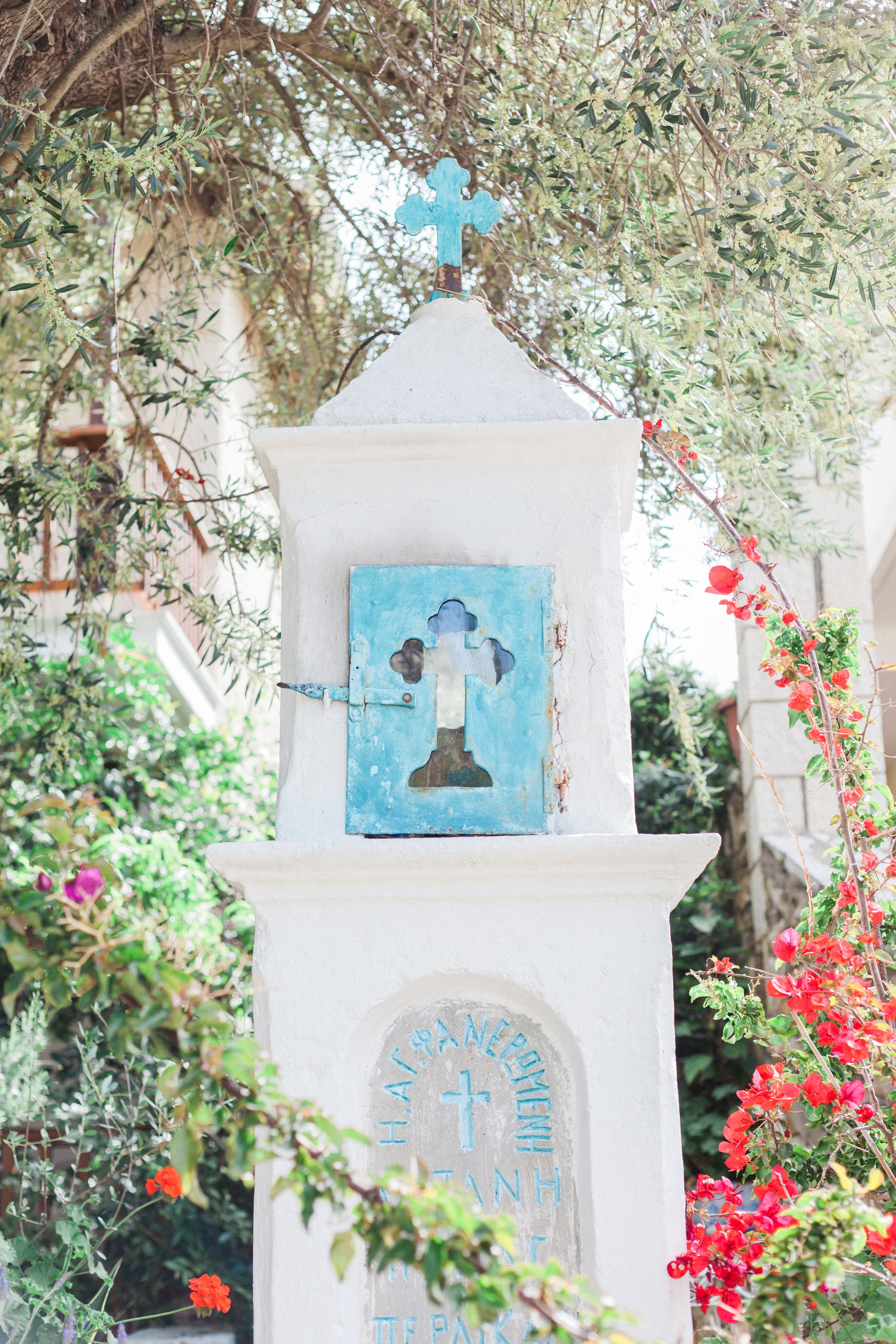 Een wit stenen monument met een blauw plaquette en een kruis bovenop, omringd door groen.
